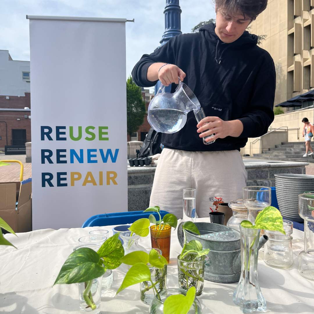A student standing by a table of plants with a "reduce renew refrain" poster behind them